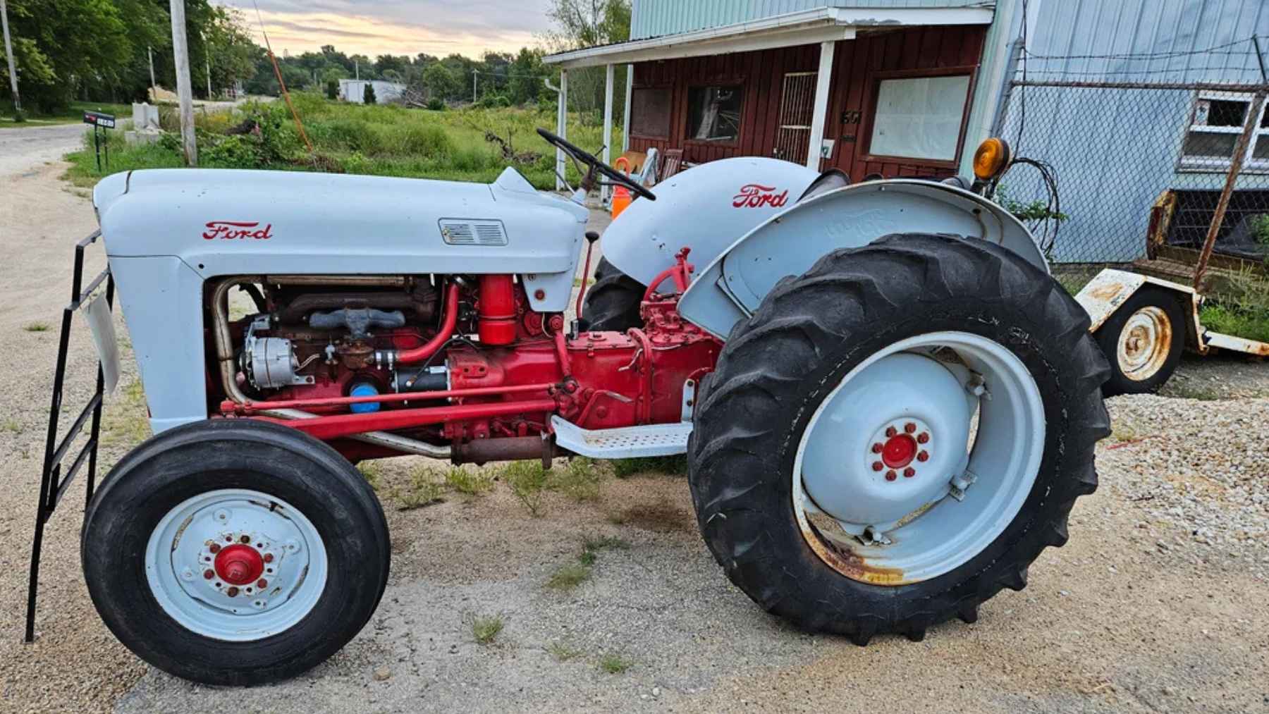 A side profile of a restored 1957 Ford 640 utility tractor with a white hood and red chassis, parked on a gravel driveway in front of a farm building.