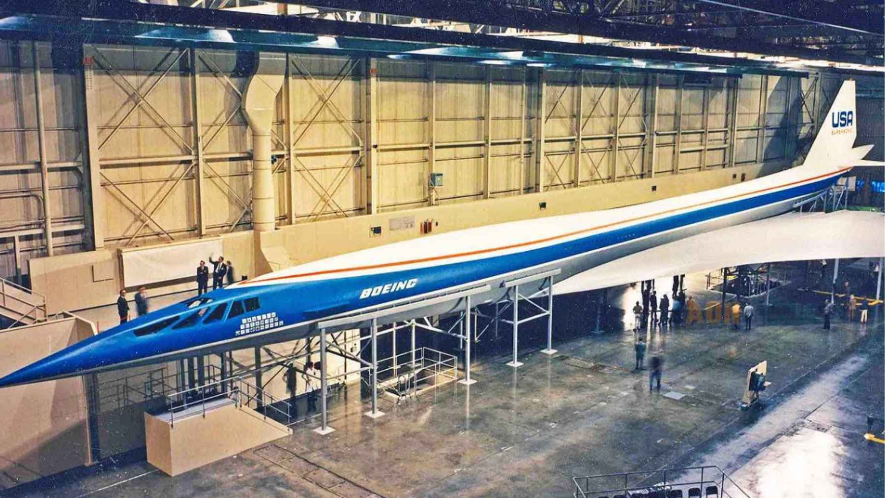 A high-angle photo looking down at the full-scale metal mock-up of the massive Boeing 2707 supersonic transport inside a large assembly hangar.