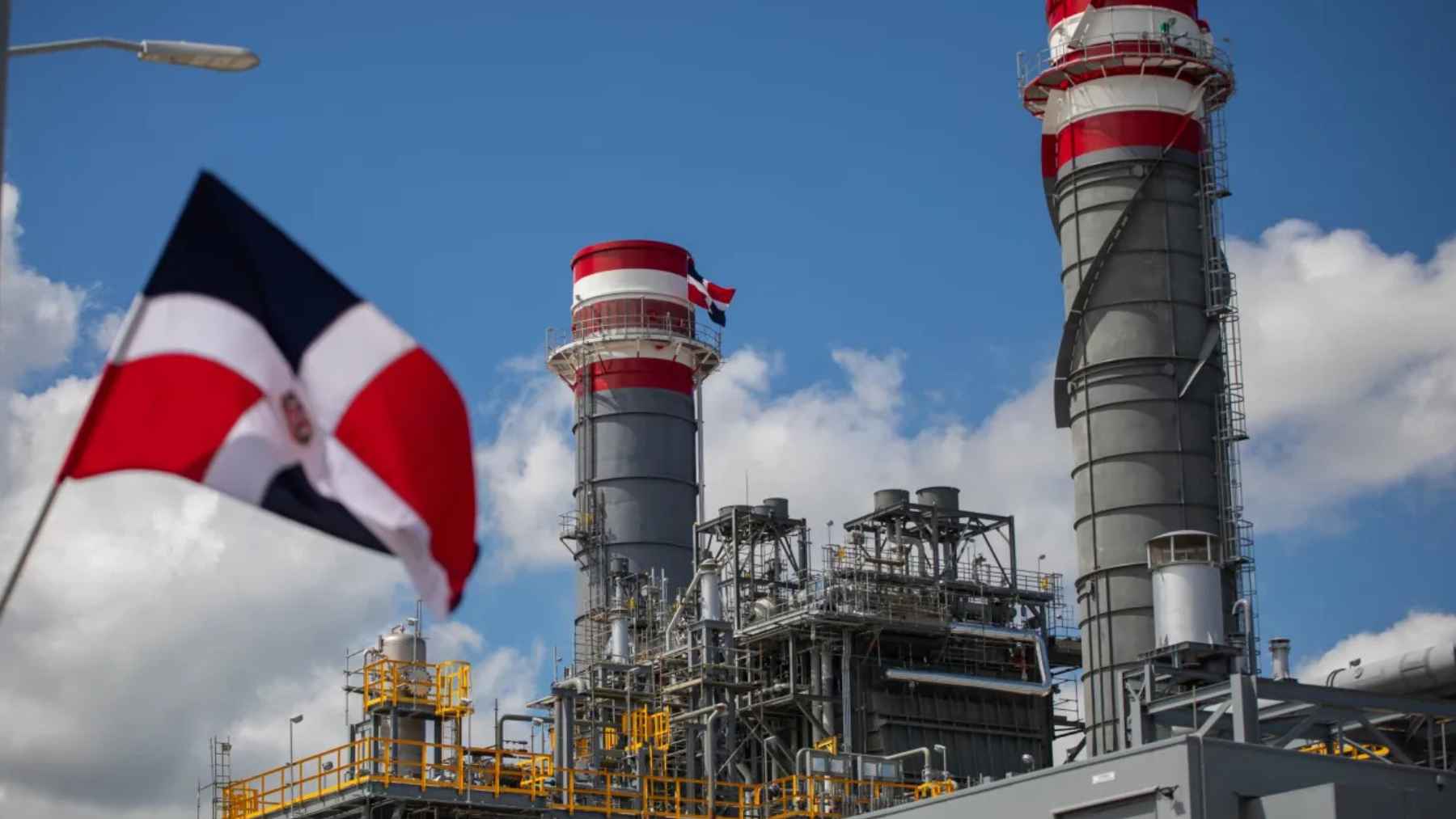 The Dominican Republic flag flying in the foreground, with the large stacks of the new 414 MW Manzanillo Power Land gas plant in the background under a blue sky.