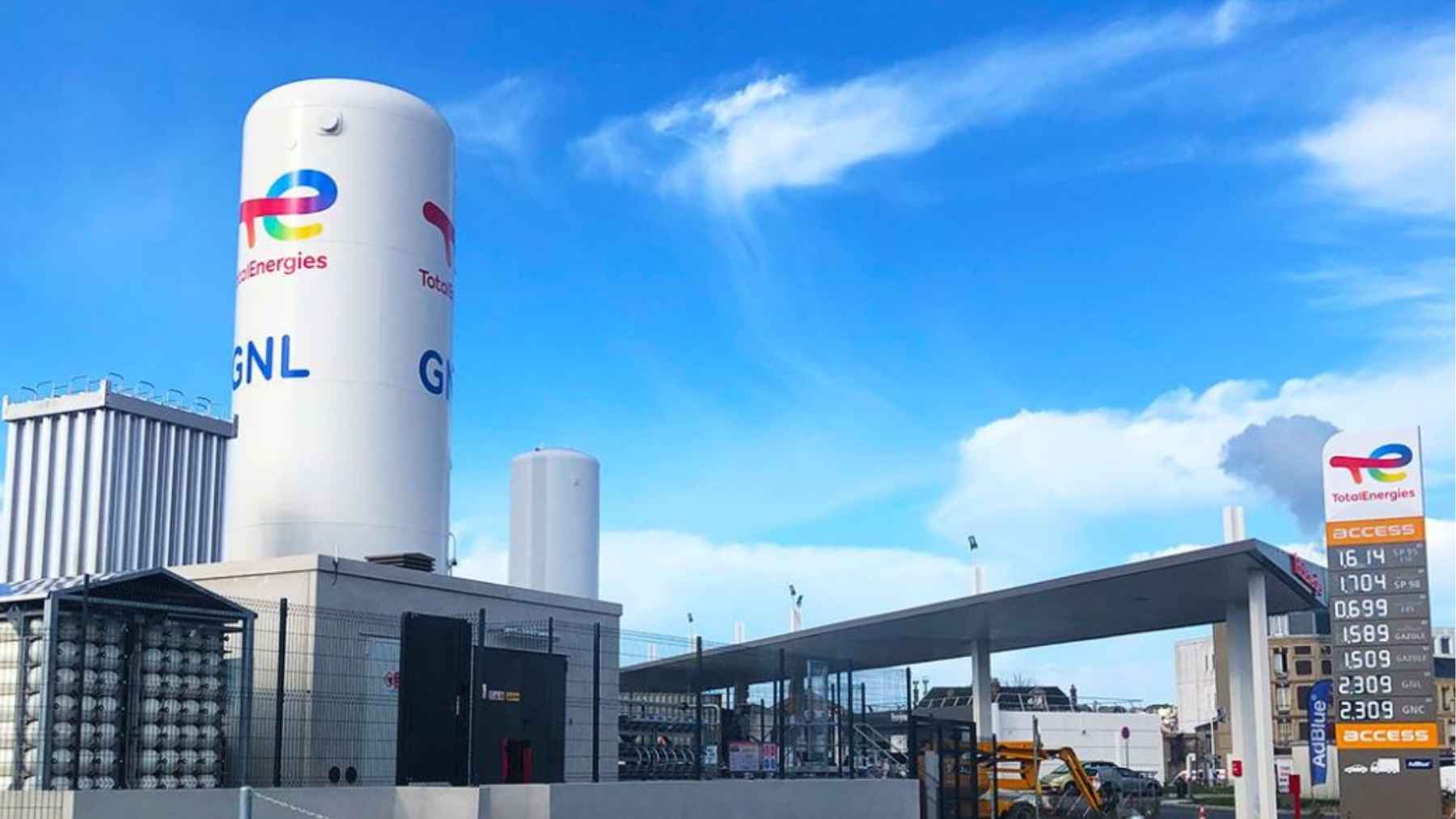 A large white TotalEnergies storage tank labeled "GNL" (LNG) next to a gas station price board under a bright blue sky.
