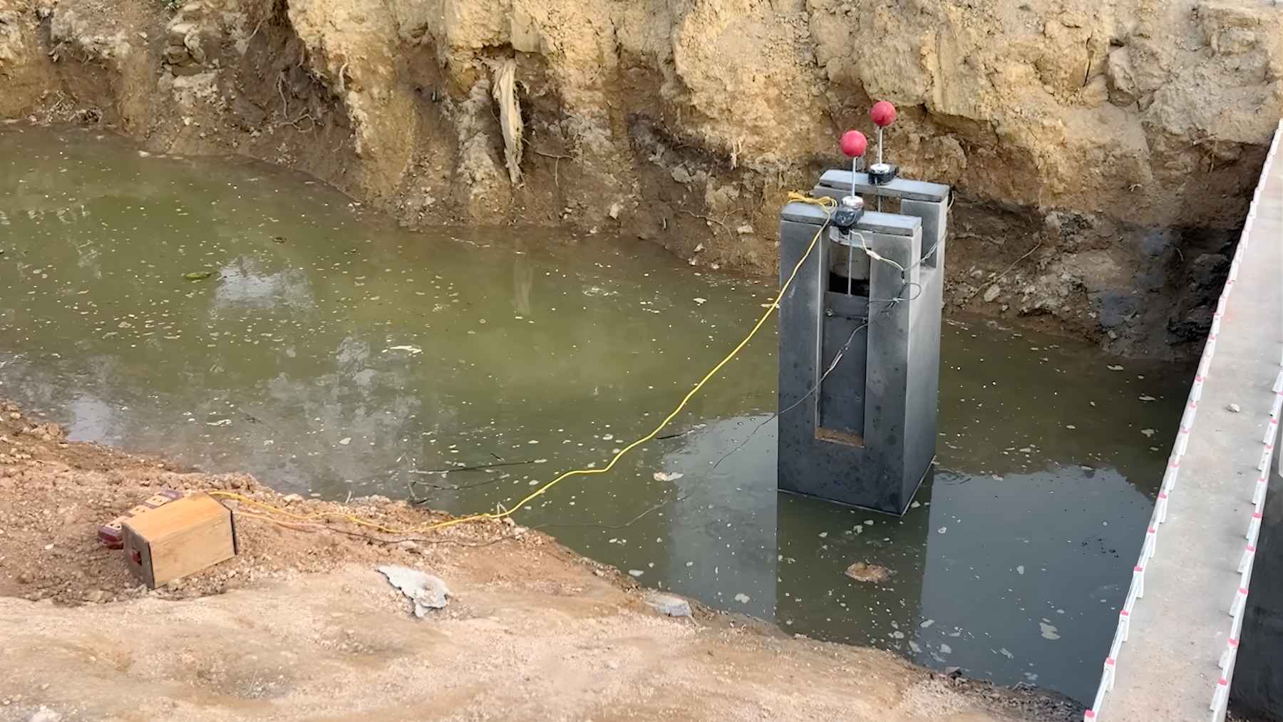 A homemade grey concrete micro-hydropower turbine structure with red handles sitting in a shallow stream in a backyard.