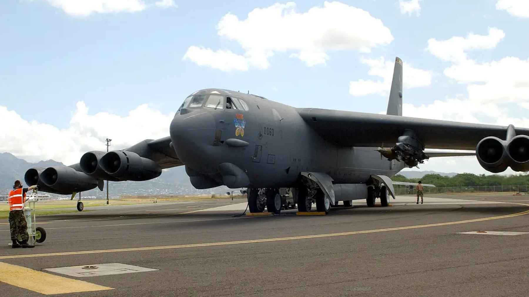 A high-resolution photo of a U.S. Air Force B-52 Stratofortress bomber on a taxiway with a ground crew member near the engines, showing a loaded weapons pylon under the wing.