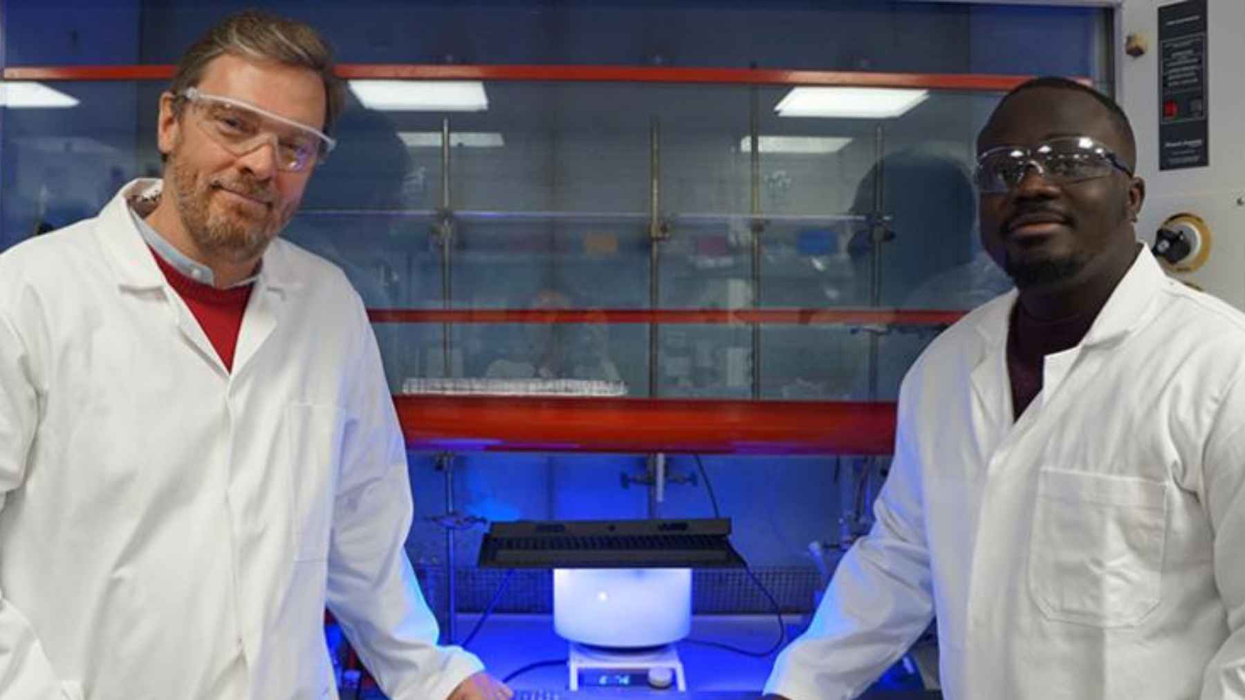 Two scientists in a laboratory standing beside a reactor used to convert battery acid and plastic waste into hydrogen