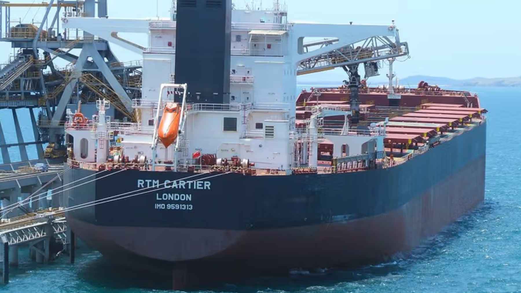 The bulk carrier RTM Cartier docked at a terminal, showing its stern and the loading infrastructure used for SimFer iron ore shipments.