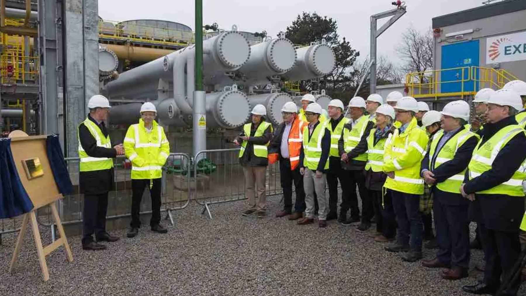 Officials and engineers in safety gear attend the inauguration of a geothermal plant in Cornwall with industrial pipes in the background.