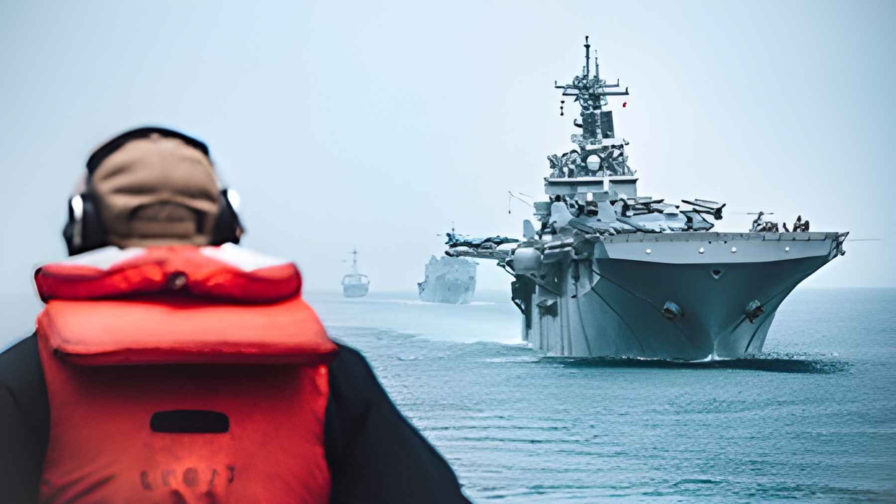 Naval vessel approaching while a crew member in safety gear observes from foreground during operations