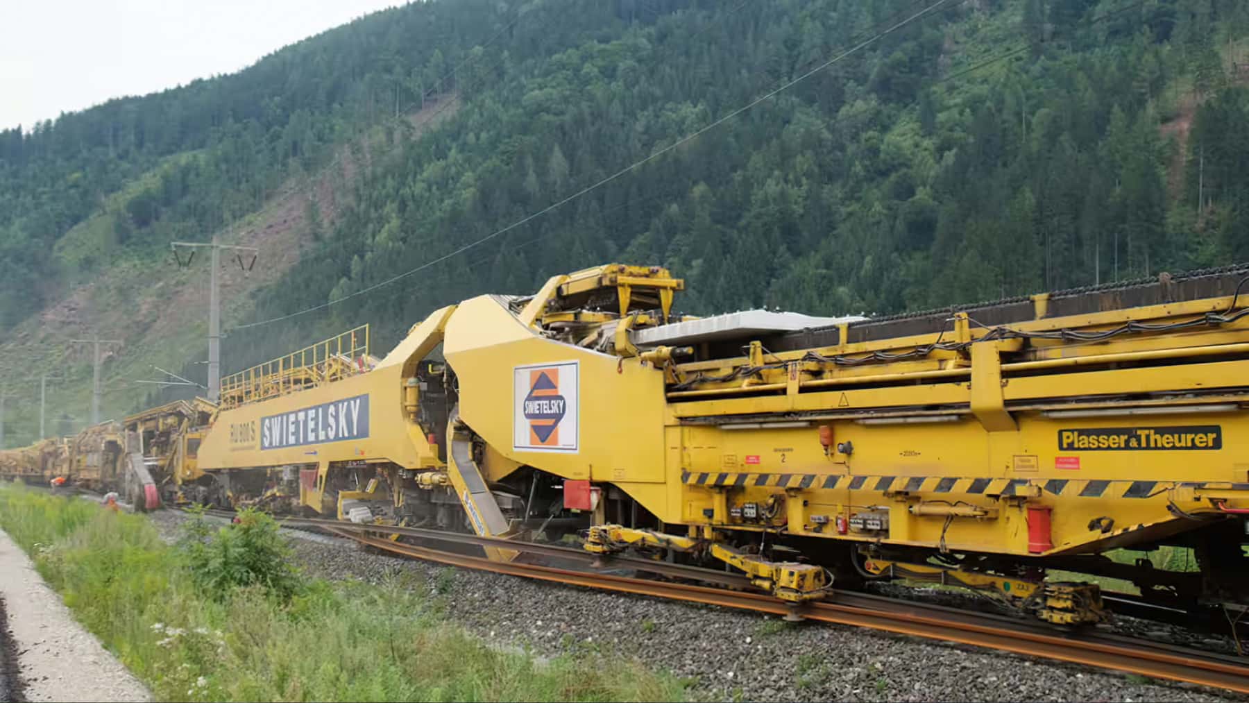 Máquina de renovación y mantenimiento de vías ferroviarias de Swietelsky y Plasser & Theurer trabajando en una línea de tren junto a una zona montañosa y boscosa