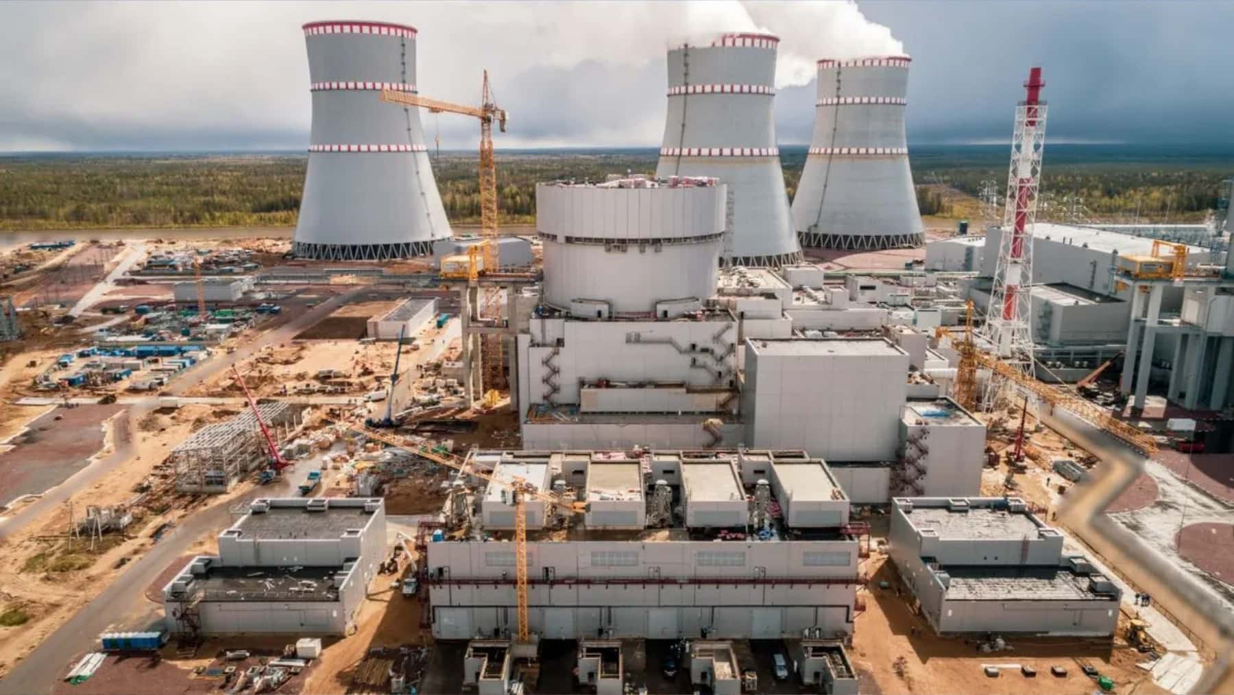 Central nuclear en construcción con edificio del reactor, grúas industriales y torres de refrigeración al fondo.