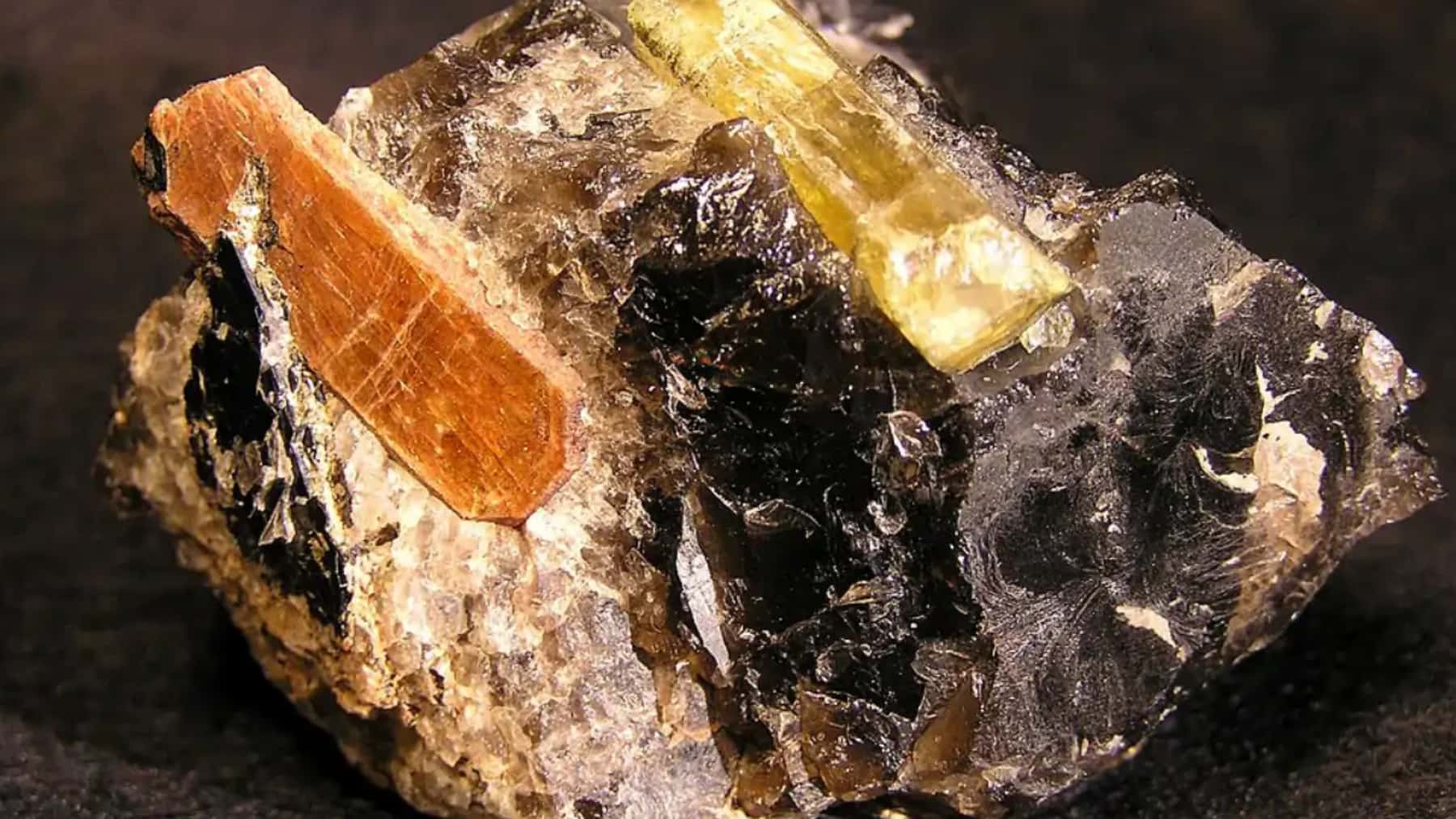 Cristal de monacita incrustado en una roca junto a otros minerales oscuros y translúcidos.