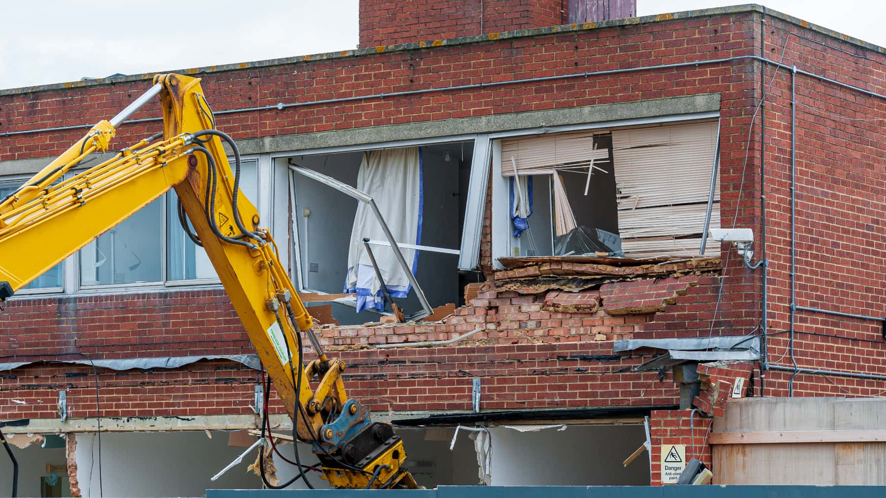 Grúa excavadora demoliendo la fachada de una vivienda de ladrillo con ventanas rotas.