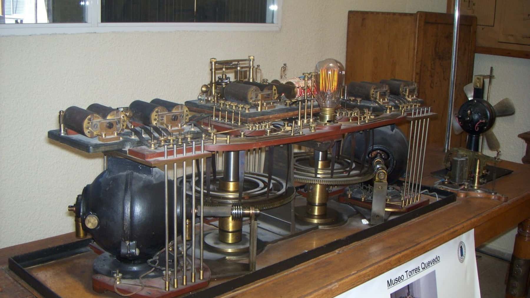 Máquina electromecánica de Torres Quevedo expuesta en un museo, con engranajes, cilindros y una bombilla encendida sobre una mesa de madera.