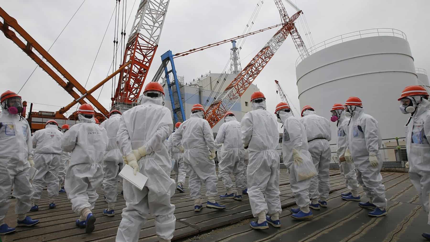 Grupo de operarios con trajes blancos, cascos naranjas y mascarillas camina en una central nuclear en Japón junto a grúas y depósitos industriales.