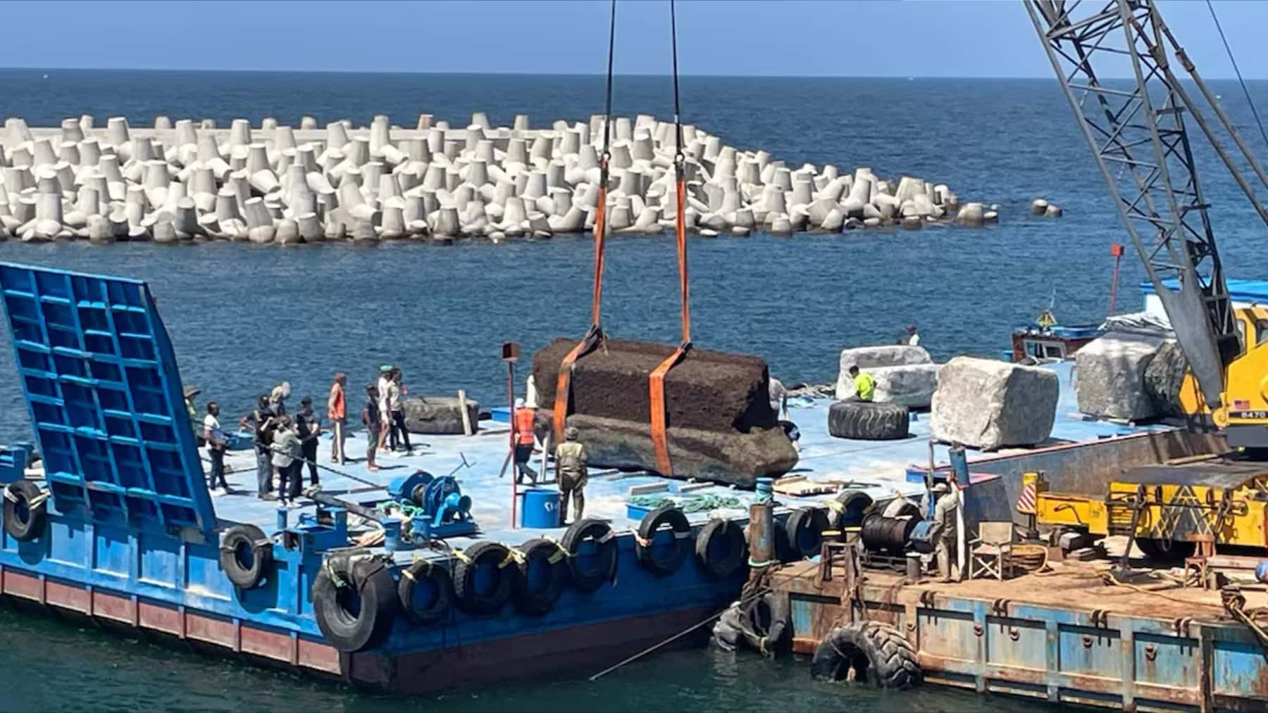 Grúa portuaria elevando un gran bloque sobre una barcaza junto a un muelle y un dique de hormigón en el mar