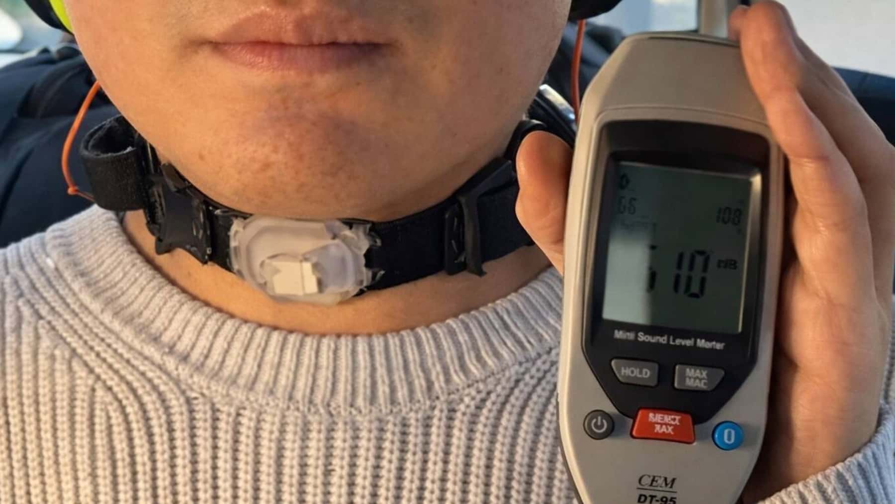 Persona con un collar en el cuello mientras sostiene un sonómetro que marca 110 dB durante una prueba de ruido.