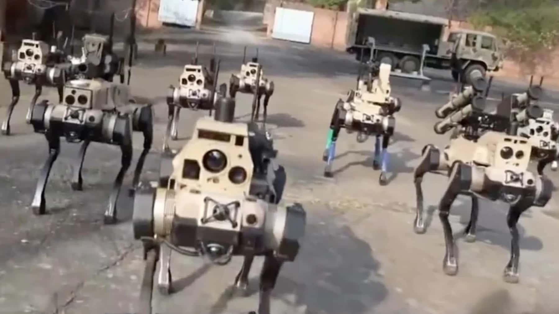 Varios perros robot de uso militar avanzan en formación sobre una explanada, con un camión al fondo.